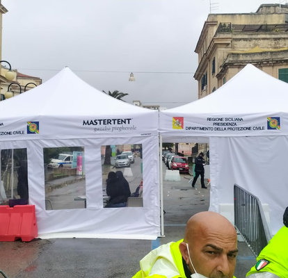 Two white folding gazebos are standing next to each other on a street, they have white sidewalls with doors and windows, in the background there are buildings and in the foreground two civil servants wearing yellow vests.