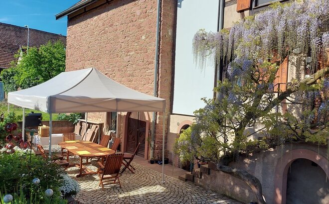 Das Gartenzelt 6x3 m in beige/ecru steht auf der Terrasse im Garten. Darunter steht eine Sitzecke mit einem großen Tisch und bequemen Stühlen. 