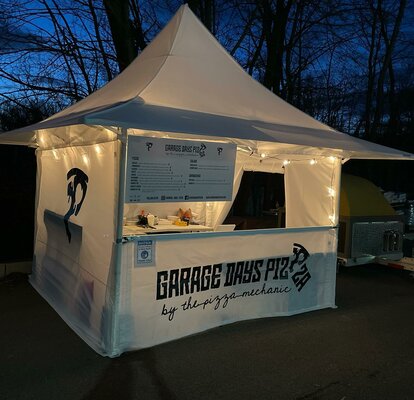 Der weiße Faltpavillon mit Vordach von "Garage Days Pizza" steht am Abend auf dem Platz. Der Street Food Stand ist mit Lichterketten dekoriert.