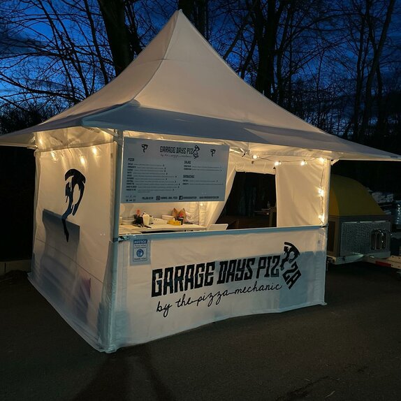 Der weiße Faltpavillon mit Vordach von "Garage Days Pizza" steht am Abend auf dem Platz. Der Street Food Stand ist mit Lichterketten dekoriert.