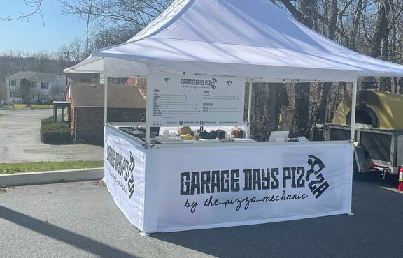 The street food stand of "Garage Days Pizza" is white and has awnings. It is printed and stands in a square.