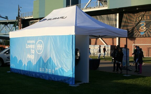 C'è un gazebo pieghevole di Subaru con tetto bianco e parete laterale azzurra su un prato. Nel fondo c'è gente.