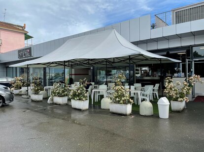 La glorieta gris claro de 8 x 4 m con 4 toldos sirve como toldo para la terraza del bar Mar'n'go en Italia. Debajo hay mesas y sillas.