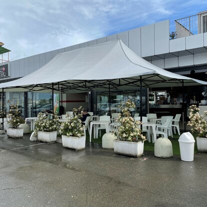 La glorieta gris claro de 8 x 4 m con 4 toldos sirve como toldo para la terraza del bar Mar'n'go en Italia. Debajo hay mesas y sillas.