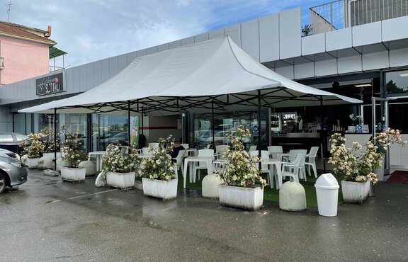 The light grey gazebo 8x4 m with 4 canopies serves as a terrace canopy for the Mar'n'go bar in Italy. There are tables and chairs underneath.