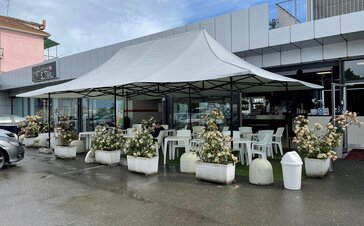 Il gazebo pieghevole grigio chiaro 8x4 m con 4 tettoie serve come copertura della terrazza del bar Mar'n'go in Italia. Tavoli e sedie sono collocati sotto.