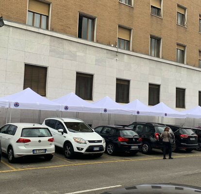 There are nine gazebos with white roofs in front of a building. There are cars in front of the gazebos and four people can be seen.