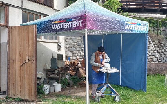 Un gazebo pieghevole con un tetto colorato e una parete laterale blu si trova nel prato di fronte a una casa. Sotto la tenda c'è una persona che sta lavorando.