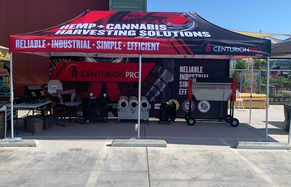A folding gazebo with a fully printed roof is standing in front of a building. Underneath the folding gazebo there are various products and a table.