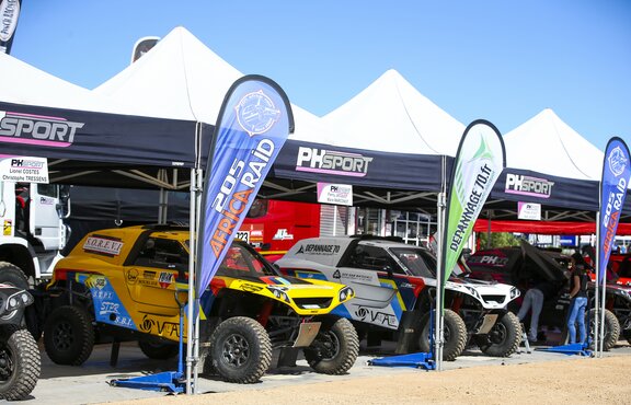Four folding gazebos with white roof and blue valance are standing next to each other. There is a race car under each of them. In front of every tent there is a printed flag.