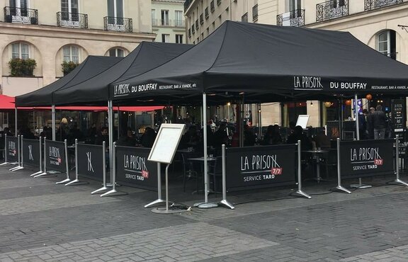 On the outdoor area of a restaurant there are standing three big, black folding gazebos with the La Prison label. Under the tents there are many tables and chairs.