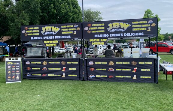 Two black, printed folding gazebos are standing next to each other on a lawn. They're stands where food and drinks are sold.