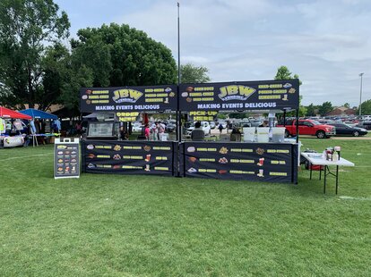Dos gazebos plegables negros y estampados están sobre un césped, uno al lado del otro. Son dos puestos donde se venden bebidas y comida.