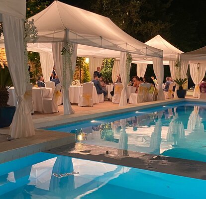 Das weiße Hochzeitszelt steht am Pool - es ist Abend. Das Hochzeitszelt ist mit tollen Beleuchtungen ausgestattet.