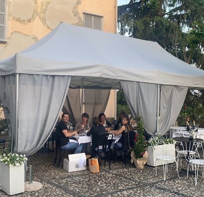 On the terrace of a restaurant there is a folding gazebo with grey roof and grey corner curtains. Underneath there are tables, chairs and guests.