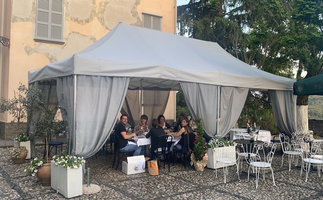 On the terrace of a restaurant there is a folding gazebo with grey roof and grey corner curtains. Underneath there are tables, chairs and guests.