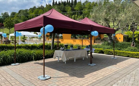 C'è un gavebo pieghevole da giardino. Sotto il gazebo ci sono dei tavoli I gazebi sono stabilizzati con dei pesi.