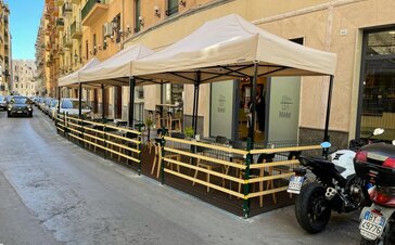 There is a tent next to the street. Undernetah it there are some tables and chairs. There ae some cars and motorcycles. Behind it there is the restaurant.