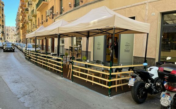 Ci sono delle tende vicino ad una strada. Ci sono delle macchine e delle moto. Sotto le tende ci sono tavoli e sedie. Dietro c'è il ristorante.