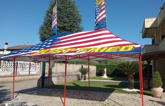 There is a gazebo in front of a garden. It has a personalized full surface print. There is street nearby and a house in the background.