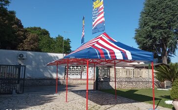 There is a folding gazebo in te backyard of a house. There is a street. The tend has two flags on its roof.