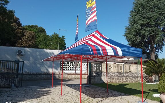 There is a folding gazebo in te backyard of a house. There is a street. The tend has two flags on its roof.