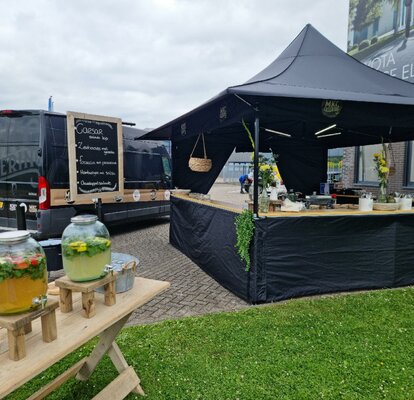 The 3x3 , tent is standing in a parking lot. The tent is black and has awnings. There are some drink containers and on the counter of the tent there are some plants and objets. 