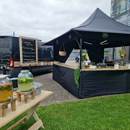 La tenda 3x3 m è in un parcheggio. La tenda è nera e ha delle tettoie. Ci sono alcuni contenitori di bevande e sul bancone della tenda ci sono alcune piante e oggetti. 