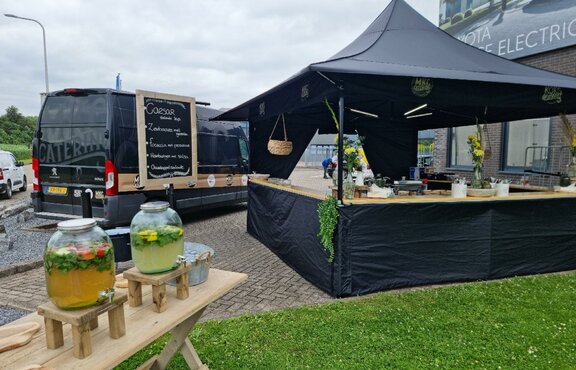 The 3x3 , tent is standing in a parking lot. The tent is black and has awnings. There are some drink containers and on the counter of the tent there are some plants and objets. 