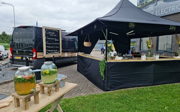 The 3x3 , tent is standing in a parking lot. The tent is black and has awnings. There are some drink containers and on the counter of the tent there are some plants and objets. 