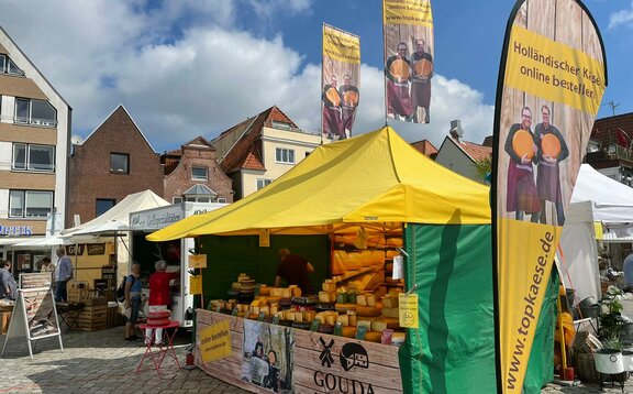 Das gelbe Marktzelt 4,5x3 m hat ein Vordach und grüne Seitenwände. Darunter wird Gouda Käse verkauft. Runderhum und auf dem Dach sind bedruckte Fahnen.