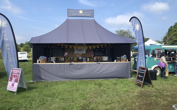 On a meadow there is a big, black folding gazebo with awning. On both sides there is a printed flag. The folding gazebo is a drinks stand.