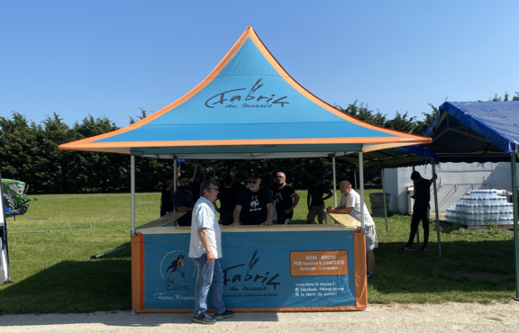There is a 3x3m folding gazebo with awnings and half-hight sidewalls.It has corners too. The roof has a personalised printing which is blue with orange elements. It is standing in a garden and there are some people behind and in front of the gazebo.