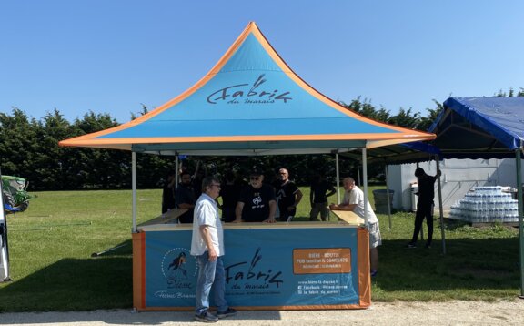 Es handelt sich um einen 3x3m großen Faltpavillon mit Vordach und halbhohen Seitenwänden, der auch Ecken hat. Das Dach hat einen personalisierten Druck, der blau mit orangefarbenen Elementen ist. Es steht in einem Garten und es gibt einige Leute hinter und vor dem Pavillon.