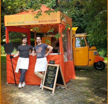 An orange 2x2 folding gazebo is printed with the company name. The gazebo has sidewalls. In front of the gazebo there are two people with kitchen wear and utensils in the hand. The gazebo is standing in front of a mobile pizza oven.  