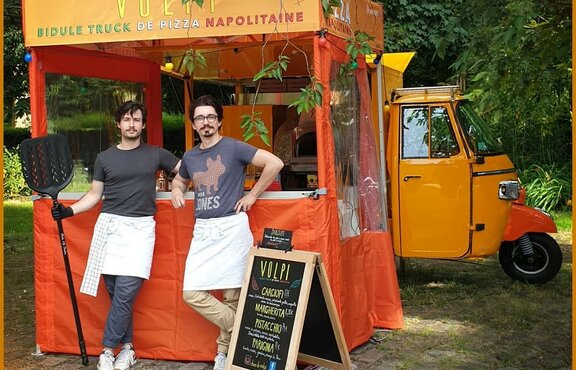 An orange 2x2 folding gazebo is printed with the company name. The gazebo has sidewalls. In front of the gazebo there are two people with kitchen wear and utensils in the hand. The gazebo is standing in front of a mobile pizza oven.  