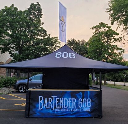 Sull'immagine c'è un gazebo nero con stampa, con un bancone in legno e una bandiera sul tetto. È un bar.
