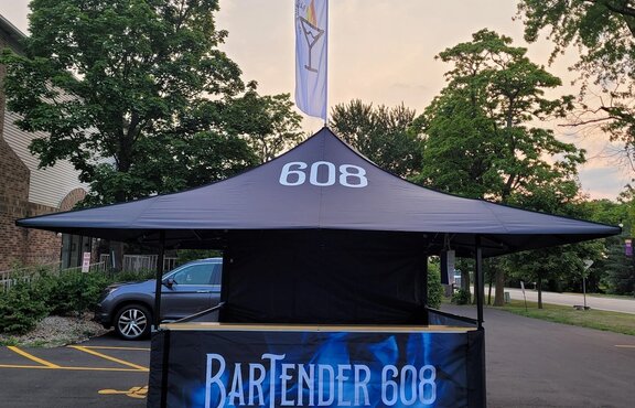 On the picture there is a black, printed folding gazebo with wooden counter and a roof-flag, It is a cocktail bar.