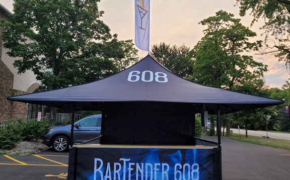 Auf dem Bild ist ein schwarzer, bedruckter Faltpavillon mit Holztheke und Dachflagge zu sehen. Er dient als Cocktail-Bar.