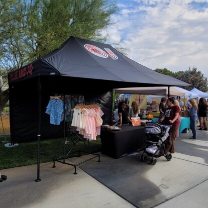 Auf einem Platz steht ein schwarzes Marktzelt mit Bedruckung und einem Vordach. Darunter hängen Kleidungsstücke.