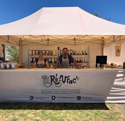 On the picture there is a folding gazebo with awning, wooden counter and printing. Underneath it there are two persons and several products. It is a folding gazebo for markets.