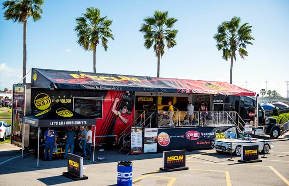 On the picture there is a big, black truck. In front of it there is a white cabrio and on its left a black folding gazebo as sales stand.