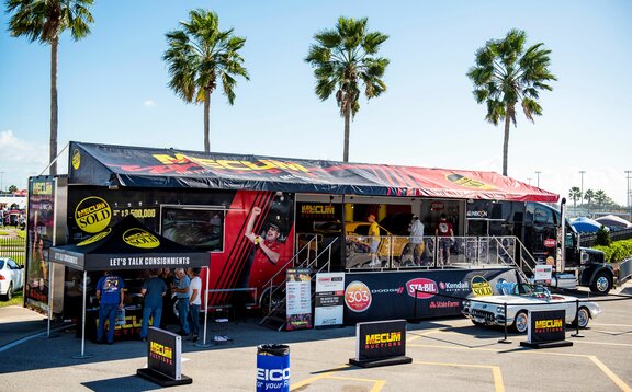 On the picture there is a big, black truck. In front of it there is a white cabrio and on its left a black folding gazebo as sales stand.