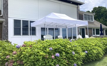 On a terrace in front of a house there is a white folding gazebo. There are some chairs under it and a hedge around the terrace.