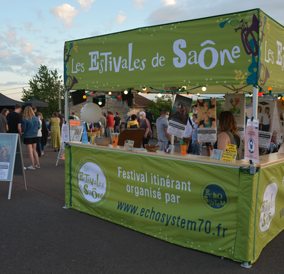 A green folding gazebo with a flat roof and a wooden counter can be seen. The tent is illuminated and people can be seen in the background.