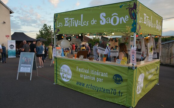 C'è un gazebo pieghevole verde con tetto piatto e un bancone di legno. Il gazebo è illuminato e sullo sfondo si vedono alcune persone.