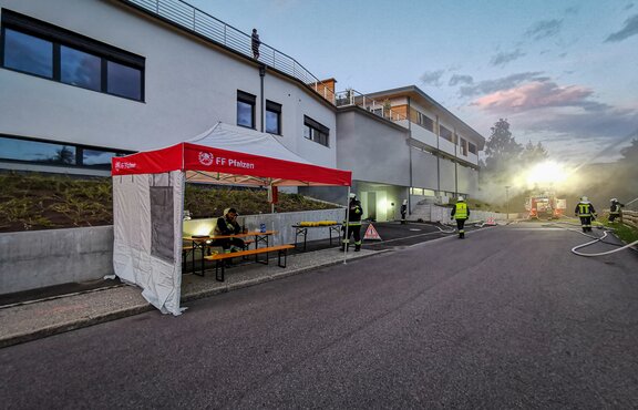 Next to a road there is a red and white fire brigade tent. Several firemen can be seen around the tent. Under the gazebo there is one fireman sitting on a bench at a table.