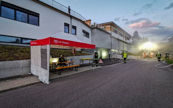 Next to a road there is a red and white fire brigade tent. Several firemen can be seen around the tent. Under the gazebo there is one fireman sitting on a bench at a table.