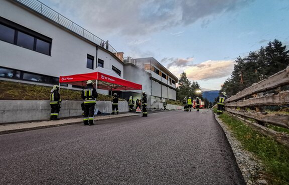 Neben einer Straße steht ein rot-weißes Feuerwehrzelt. Rund um das Zelt sind mehrere Feuerwehrmänner zu sehen.