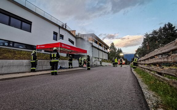 Next to a road there is a red and white fire brigade tent. Several firemen can be seen around the tent.